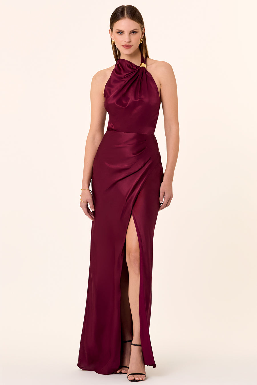 A model wearing a dark plum colored dress with a halter neckline, sleeveless design, and a high slit on the left side. The dress is made of a silky material.